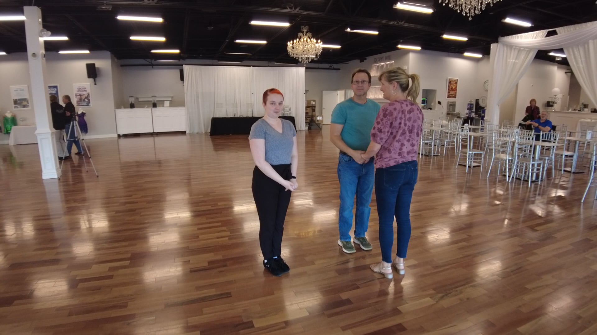 Three adults chatting on a polished wooden dance floor in a large event hall with chandeliers and white drapery.