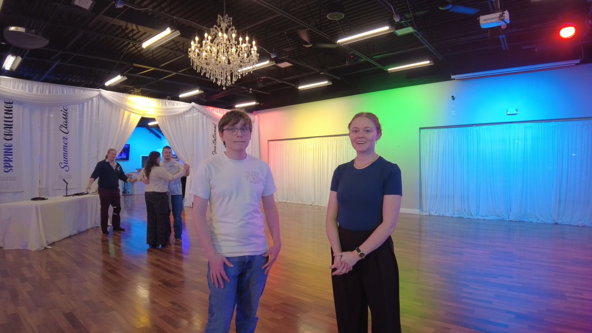 Two people stand in a dance studio with a wooden floor, draped white curtains, and rainbow-colored lighting on the back wall. A chandelier hangs from the ceiling.