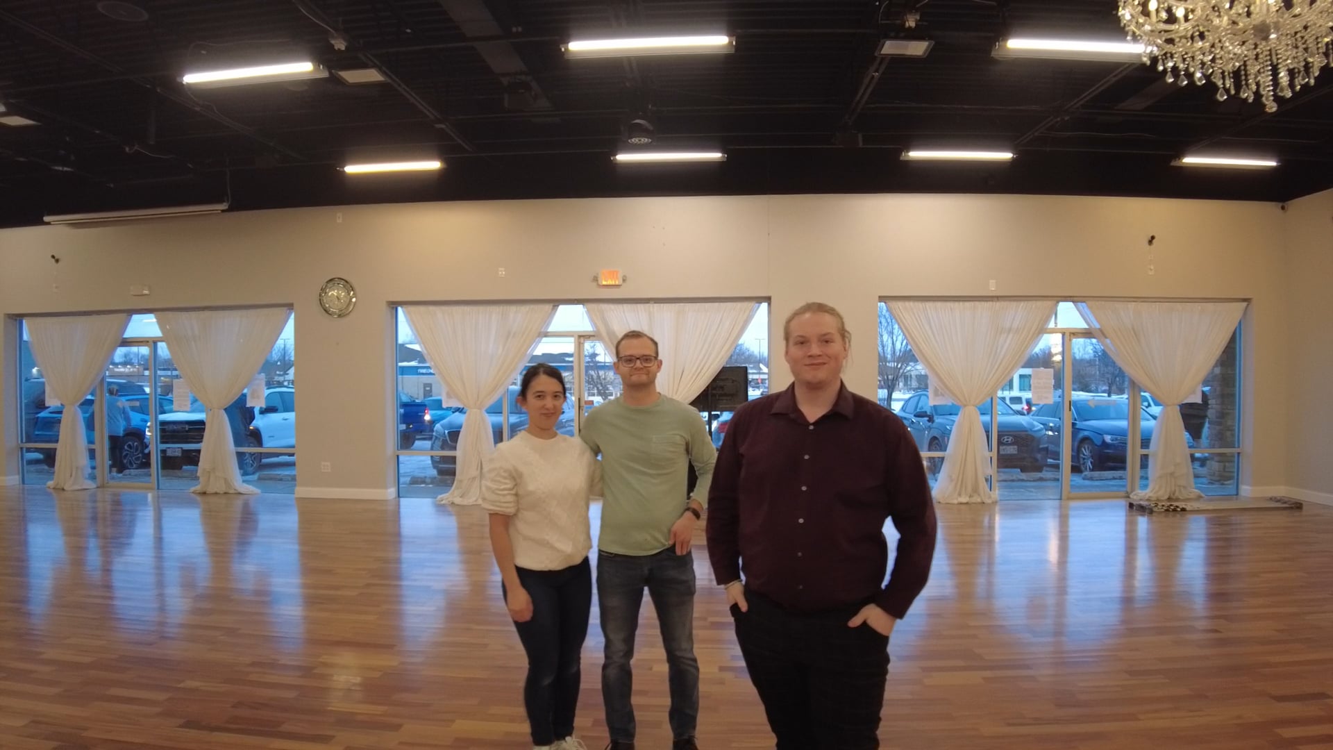 Three people stand together in a spacious dance studio with a wood floor and white curtains at the back, posing for a photo.