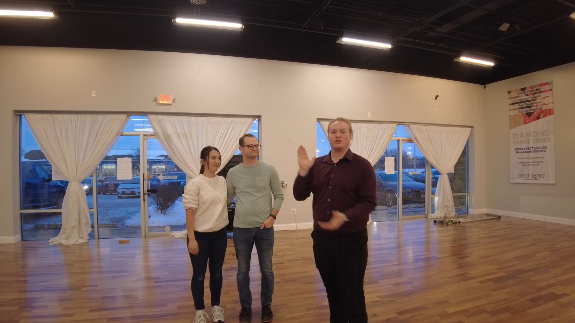 Three people stand together in a bright dance studio with wooden floors and large windows with white curtains behind them.