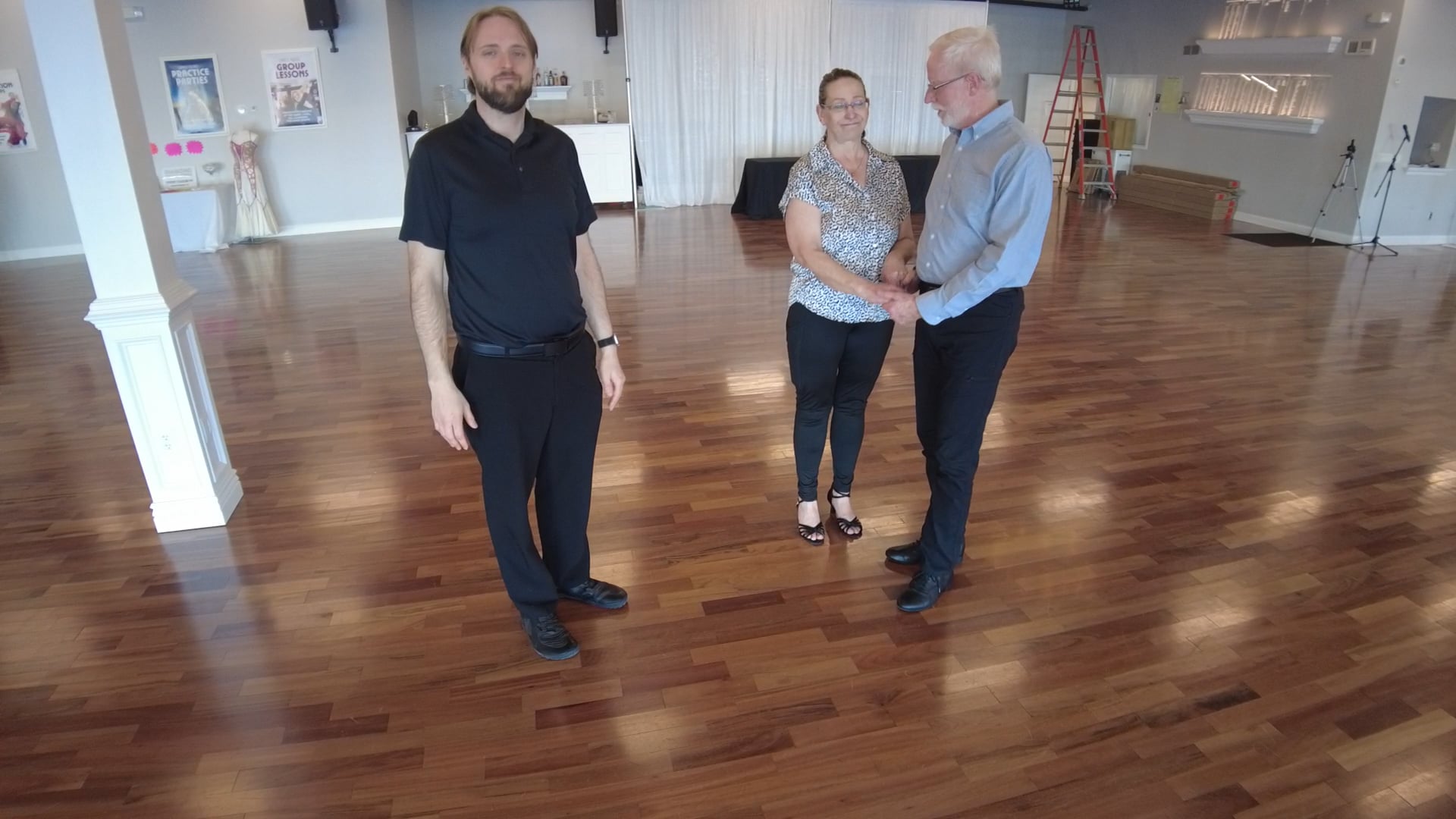 Three adults stand in a bright dance studio with a polished wood floor; a couple shakes hands while another man watches.
