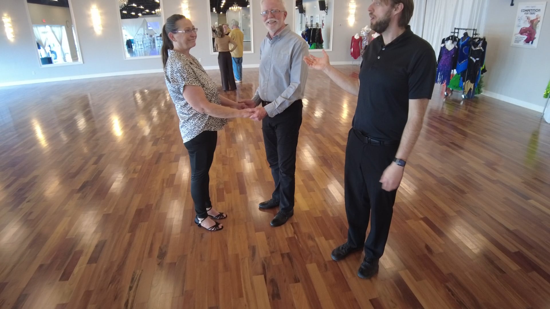Three adults stand in a dance studio forming a ballroom hold, smiling at each other; woman in patterned blouse, man in light shirt, another man in black polo nearby, with mirrors and costumes in the background.