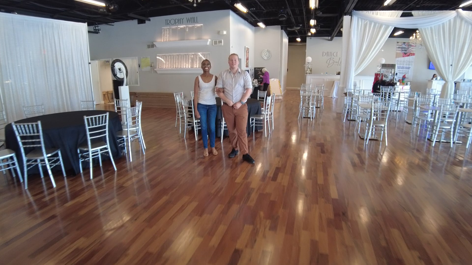Two people stand arm in arm in a bright event venue with white chairs, round tables with black cloths, and draped white fabric in the background.