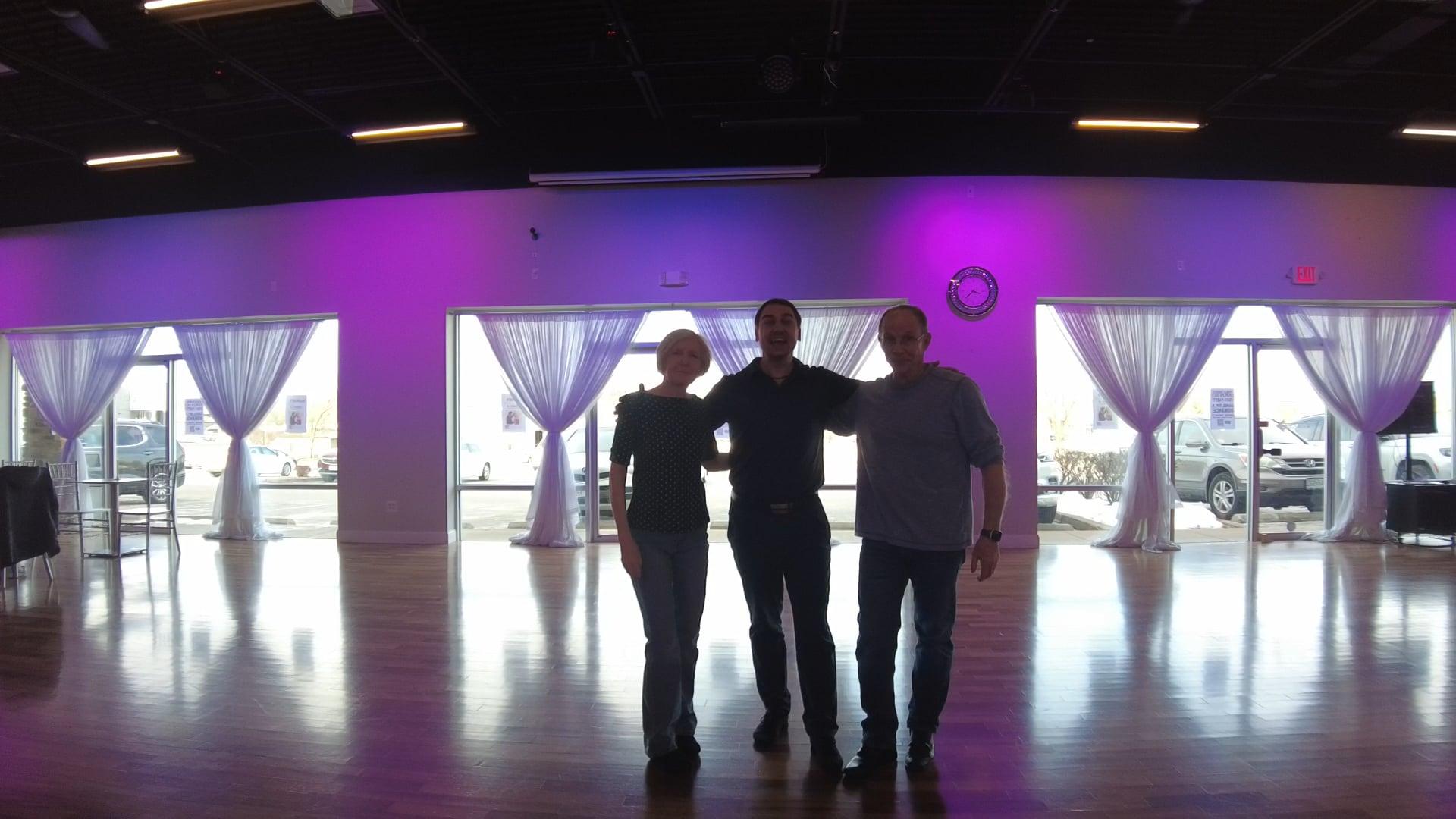 Three people pose together in a spacious dance studio with purple uplighting and sheer-curtained windows behind them.